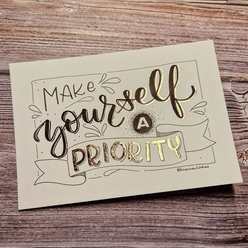 Ein Handlettering Projekt liegt auf einem Holztisch. In verschiedenen Schriftarten (Sans-Serif, Brushscript) und mit verschiedenen Schmuckelementen steht geschrieben "Make yourself a priorty". Das Lettering ist mit Goldfolie veredelt.