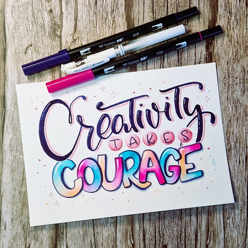 Auf einem Holztisch liegt ein Papier mit dem Schriftzug "Creativity takes courage". Darüber liegen dekorativ die zugehörigen Stifte, Tombow ABT Brushpens in den Farben pink und dunkelblau und ein weißer UniBall Signo Gelstift. Der Spruch ist im Handlettering-Stil gestaltet.