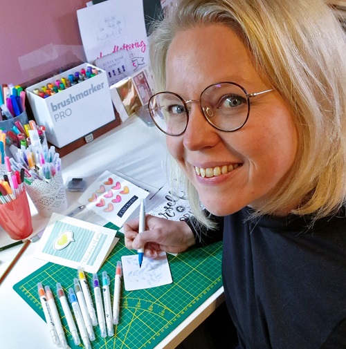 Tina sitzt an ihrem Kreativplatz und gestaltet eine Karte im Handlettering Stil. In der Hand hält sie einen Brushpen. Im Hintergrund stehen viele verschiedene Stifte.