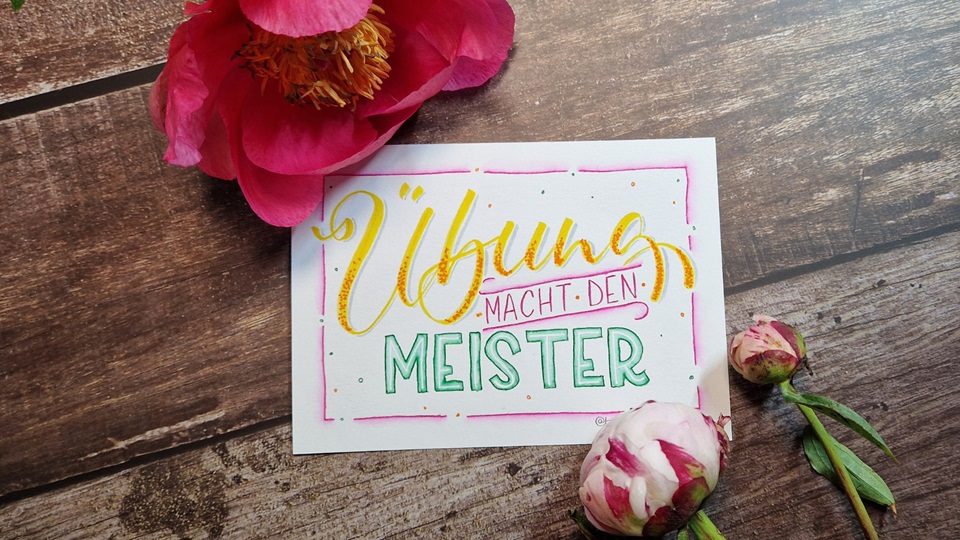 Das Bild zeigt ein farbenfrohes Handlettering mit den Worten "Übung macht den Meister" der Handlettering Künstlerin Tina Niemann. Es liegt auf einem Holztisch und ist dekoriert mit Pfingstrosen.