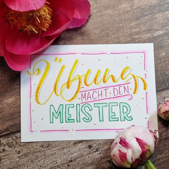 Das Bild zeigt ein farbenfrohes Handlettering mit dem Sprichwort "Übung macht den Meister" der Handlettering Künstlerin Tina Niemann. Es liegt auf einem Holztisch und ist dekoriert mit Pfingstrosen.