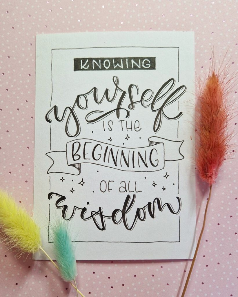 Das Bild zeigt das Ergebnis einen Handlettering-Prozesses mit dem Spruch "Knowing yourself is the beginning of all wisdom". Es ist in schwarz-weiß gehalten, liegt auf einem rosa Untergrund und ist dekoriert mit bunt eingefärbten Gräsern.