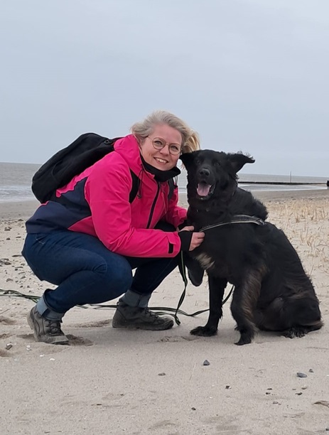 Tina hockt mit ihrem Hund am Strand. Der Wind bläst, die Ohren des Hundes flattern im Wind