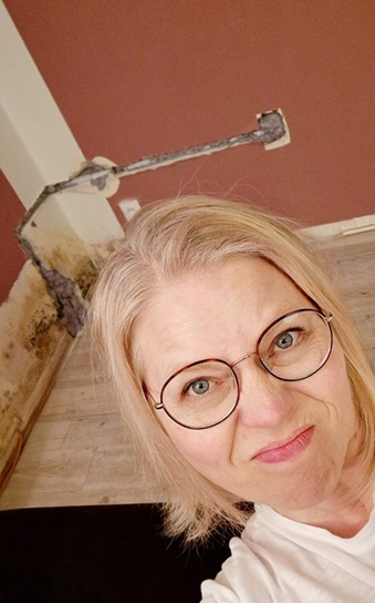 Tina sitzt vor ihrer Wand im Wohnzimmer. Zu sehen ist ein Wasserschaden, Schimmel und eine bereits aufgestemmte Wand