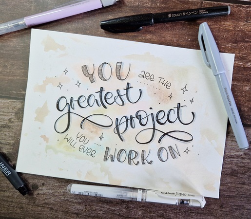 Auf einem Tisch liegt ein DIN-A5 Papier mit deinem Lettering mit einem Watercolor-Hintergrund und mit dem Schriftzug "You are the greatest project you will ever work on". Es ist umgeben von lose herumliegenden, verschiedenen Stiften.