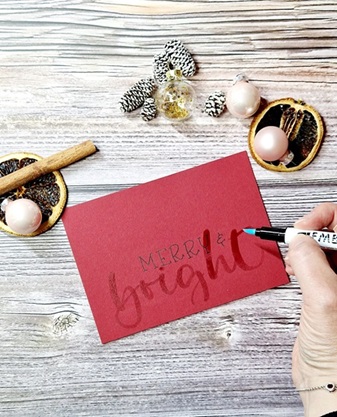 Auf einem weihnachtlich dekorierten Tisch liegt eine rote Karte, auf der in schwarz das Wort "merry" steht. MIt einem speziellen Embossing-Stift ist das Wort "bright" darauf geschrieben worden. Der Embossing-Stift schimmert noch feucht.