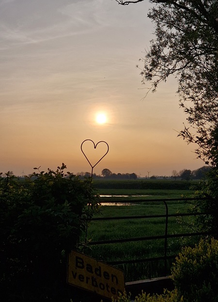 Mitten in Friesland, Ausblick über eine Wiese, die Sonne geht langsam unter, mittig im Bild steht ein dekoratives Herz.