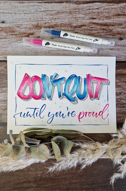 Auf dem Bild liegt ein Handlettering in pink und blau auf einem Tisch mit dem Text "Don't quit until you're proud".