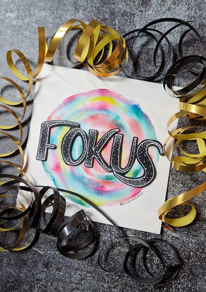 Das Bild zeigt ein Lettering mit einem farbenfrohen Hintergrund, darauf in dicken schwarzen Lettern das Wort "Fokus". Rundherum sind goldene und schwarze Luftschlangen.